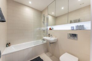 Beiges Badezimmer mit Badewanne samt Duschwand, Wand-WC, Waschbecken und Wandnische.