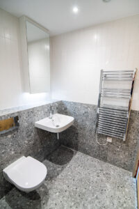 Badezimmer in grauem Terrazzo mit Wandwaschbecken, Spiegelschrank und Chrom-Heizkörper.