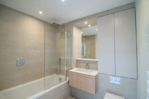 Badezimmer mit Badewanne samt Duschwand, beigen Fliesen, Holzwaschtisch und Spiegelschrank.