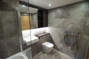 Graues Badezimmer mit Badewanne hinter Glaswand, Wand-WC und goldenem Akzent in der Nische.