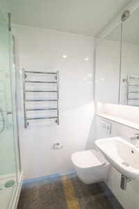 Weißes Badezimmer mit Duschkabine, Wand-WC, Waschbecken und Chrom-Heizkörper.