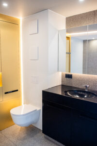 Badezimmer mit gelber Nische, schwarzem Waschtisch und Wand-WC auf Terrazzoboden.