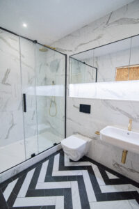Badezimmer mit Marmorfliesen, verglaster Walk-in-Dusche, goldenen Akzenten und Fischgrätboden.