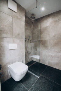 Graues Badezimmer mit Walk-in-Dusche samt Sitz, Wand-WC und dunklem Boden.