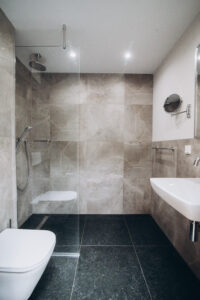 Graues Badezimmer mit Walk-in-Dusche, Wand-WC, Waschbecken und dunklem Boden.