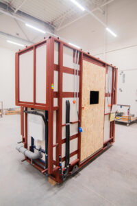 Vorgefertigtes Badmodul auf rotem Rahmen mit OSB-Tür und Sanitärinstallation.