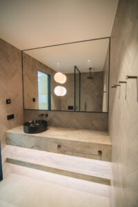 Badezimmer mit beigen Fliesen, Steinwaschtisch mit schwarzem Aufsatzbecken, großem Spiegel und Walk-in-Dusche.