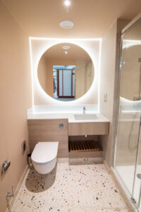 Badezimmer mit rundem beleuchtetem Spiegel, Holzwaschtisch, Terrazzoboden und Walk-in-Dusche.