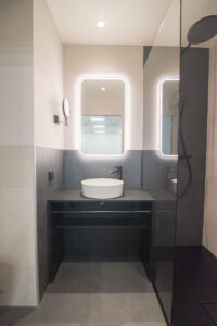 Graues Badezimmer mit beleuchteten Spiegeln, Aufsatzwaschbecken auf schwarzem Unterschrank und Walk-in-Dusche.